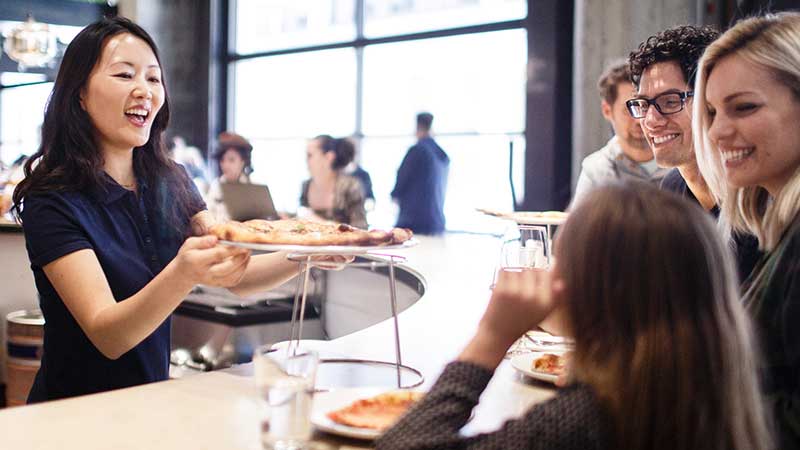At a restaurant, a young woman serves a pizza to a smiling family. At a restaurant, a young woman serves a pizza to a smiling family.
