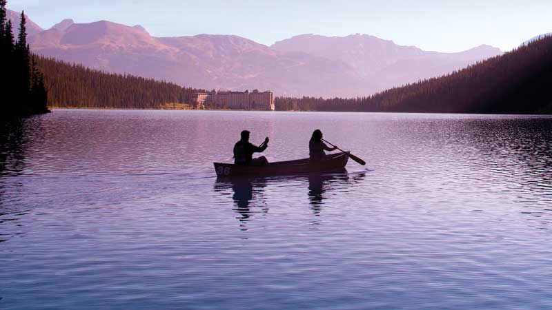 Two people in a boat paddling in a fjord during sunset with mountains, trees, and a castle in the distance.