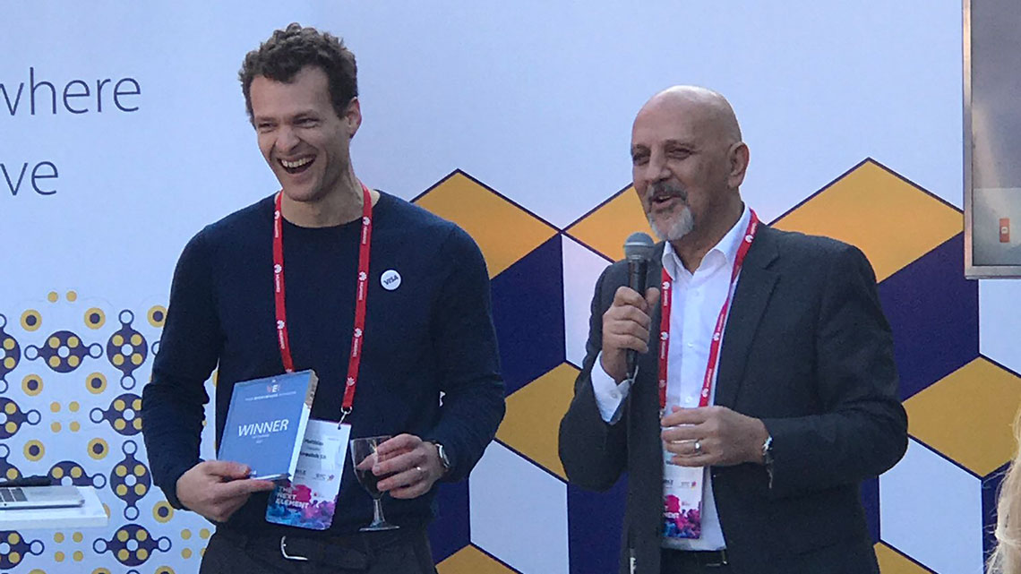 Two men standing next to each other with the man on the left holding an award and the man on the right holding a microphone.