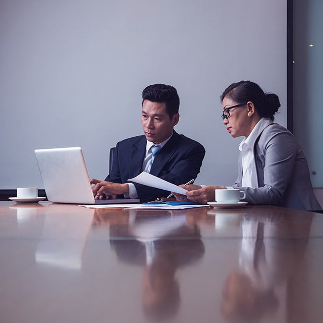 Two professionals discussing spend management while seated at a conference table with a laptop. 